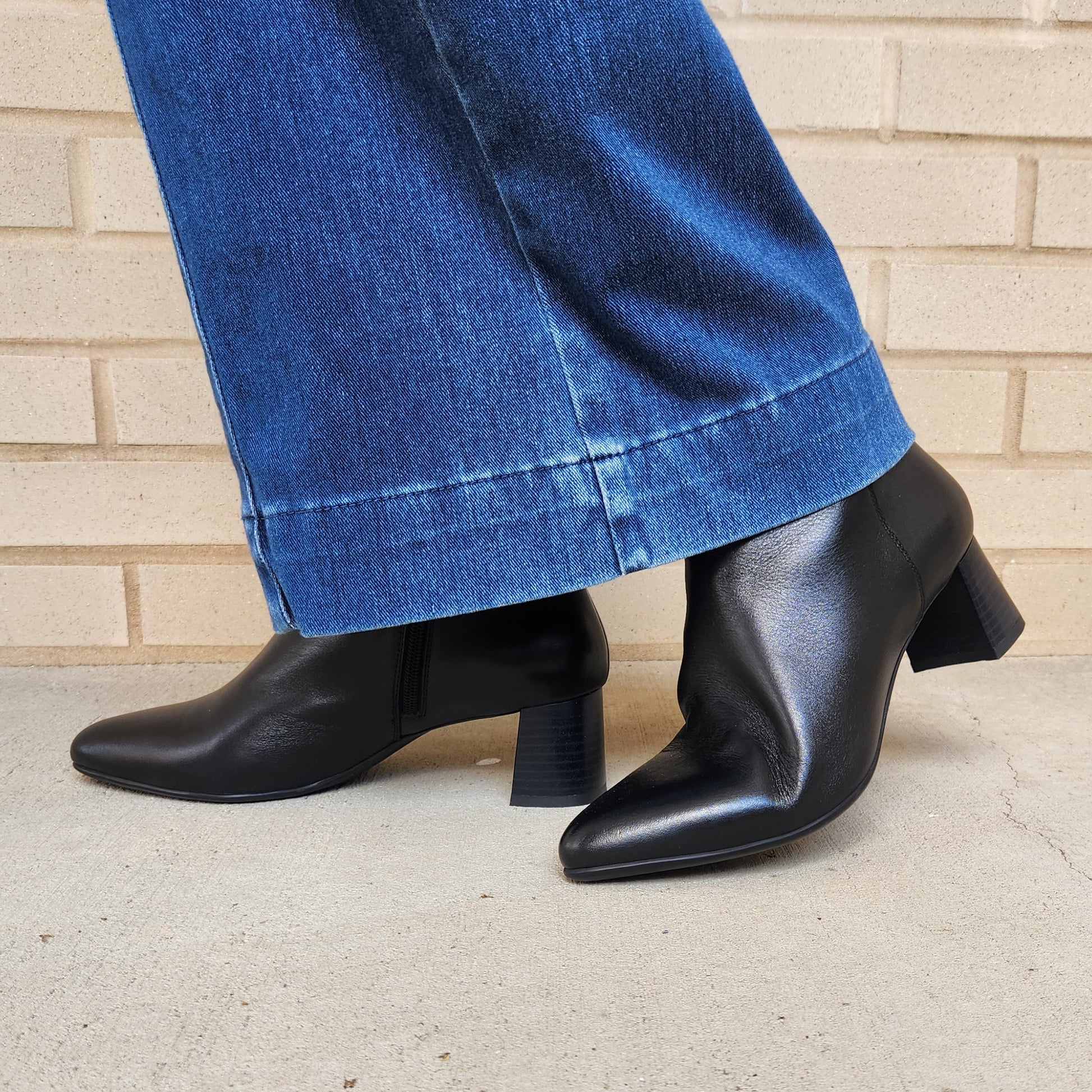 Black leather ankle boots with block heels and pointed toes, shown on a model wearing blue jeans against a light brick wall and concrete floor.