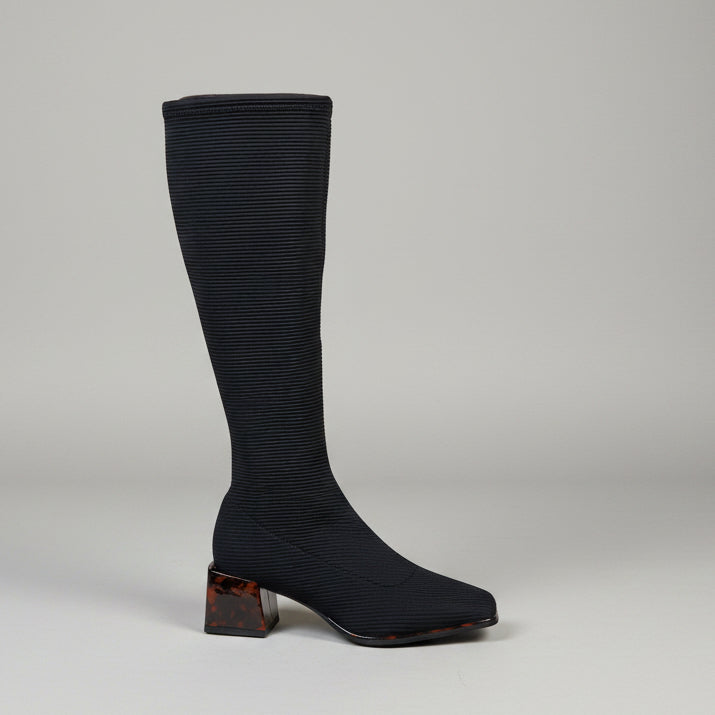 A single knee-high black ribbed boot with a square toe and a brown marbled block heel, photographed standing on a concrete floor against a light beige brick wall.