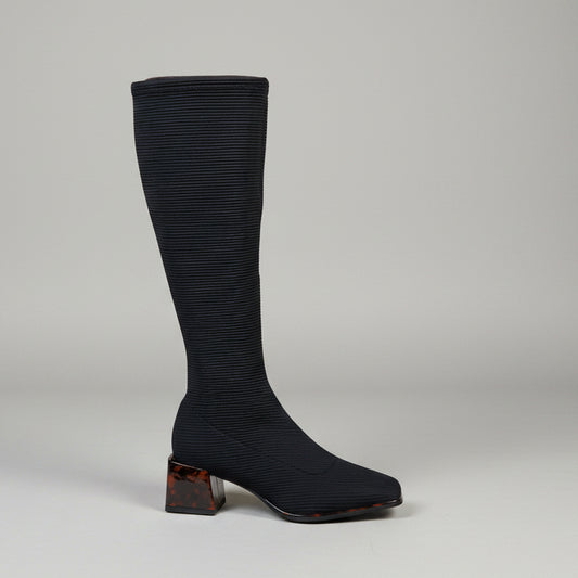 A single knee-high black ribbed boot with a square toe and a brown marbled block heel, photographed standing on a concrete floor against a light beige brick wall.