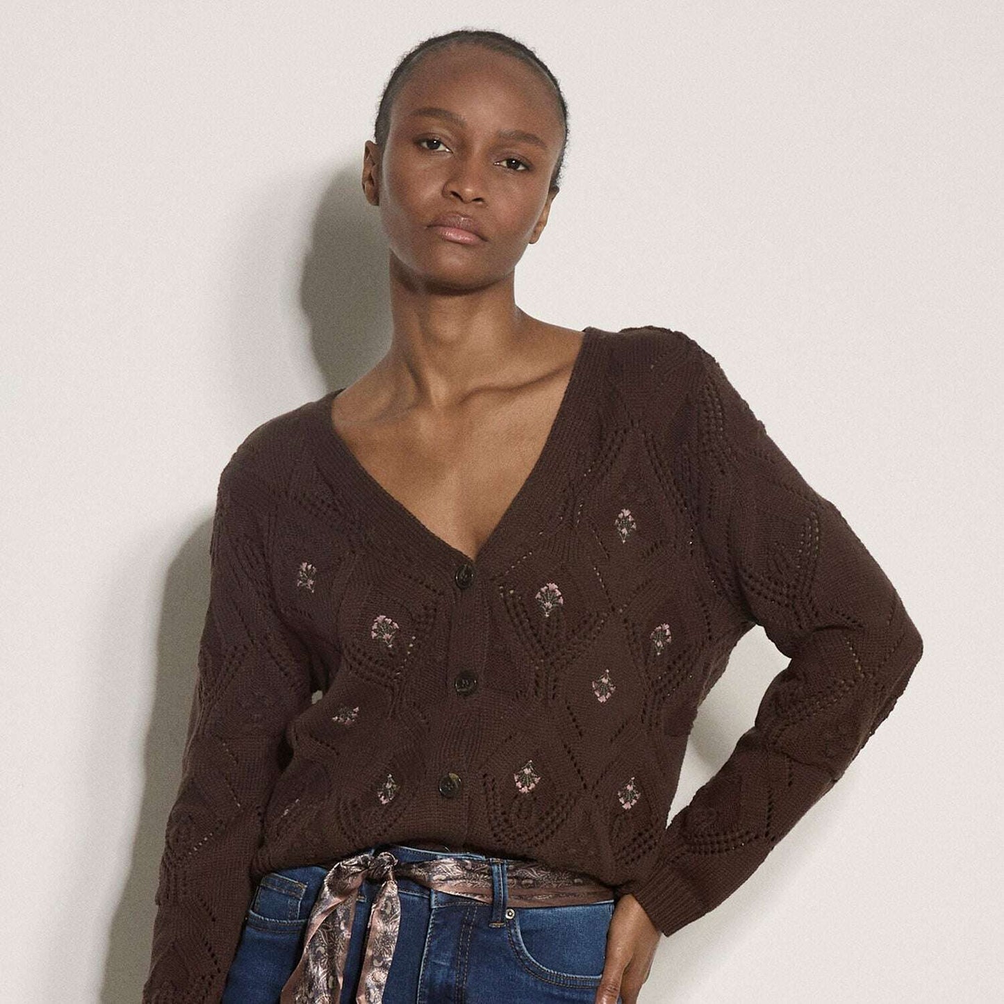 A model wearing a dark brown cable-knit cardigan sweater with decorative embroidered details, paired with blue jeans and a patterned belt, standing against a plain off-white wall.