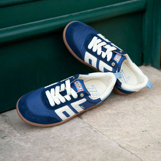A pair of blue slip-on sneakers with white graphic lettering and white laces, resting on a stone step in front of a dark green wooden door.