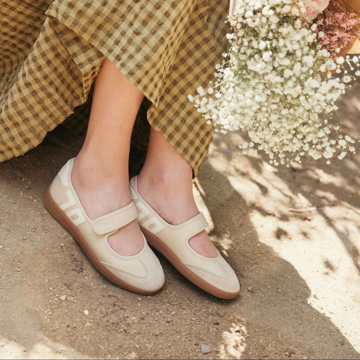 A person seated outdoors wearing beige Mary Jane-style flat shoes with a brown sole, paired with a brown and beige gingham skirt and holding a bouquet of white flowers.