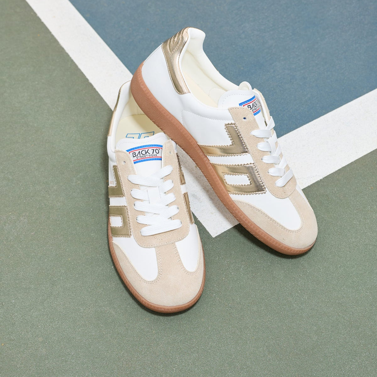 A pair of low-top sneakers in beige, white, and metallic gold, styled on a green and blue court floor.