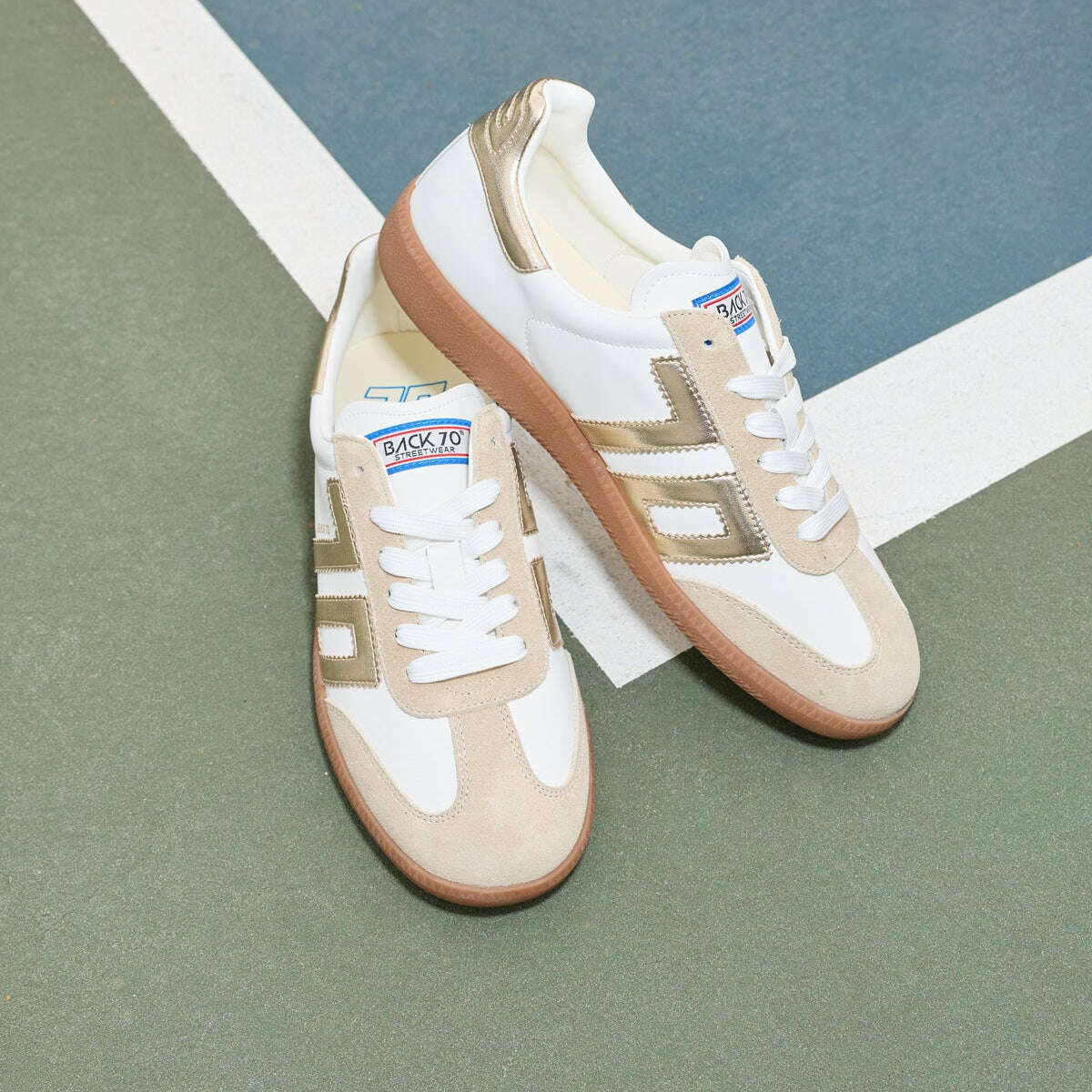 A pair of low-top sneakers in beige, white, and metallic gold, styled on a green and blue court floor.