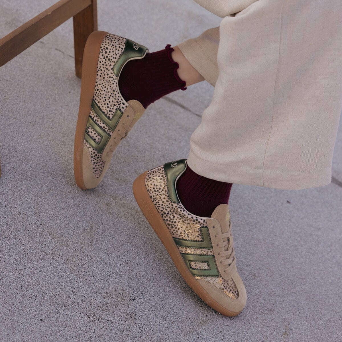 A person seated on a wooden bench wearing beige patterned sneakers with green side stripes, burgundy socks, and cream trousers, photographed outdoors on a light concrete surface.