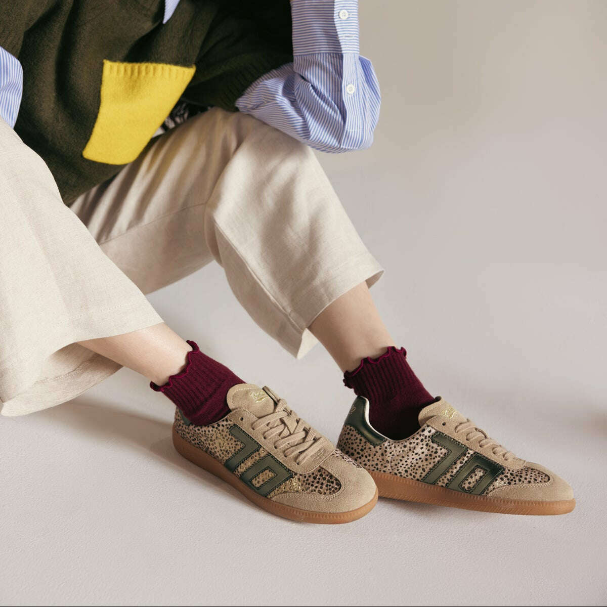 A person seated cross-legged wearing beige and olive low-top sneakers with a spotted pattern, burgundy socks, and a layered outfit against a plain light gray studio background.