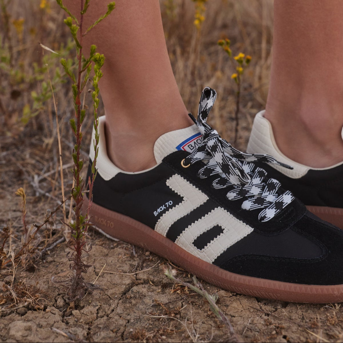 A close-up of a person's legs standing in dry grass outdoors, wearing black and off-white Samba sneakers with patterned laces and gum soles.