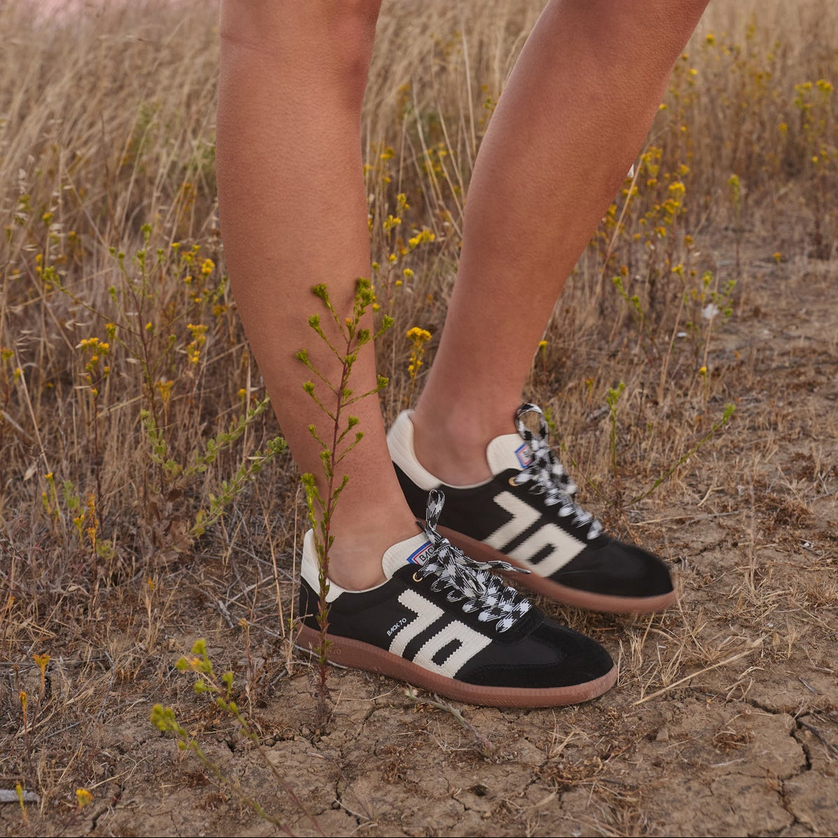 A person outdoors in a dry grassy field at sunset wearing black low-top sneakers with large white '76' lettering and white-and-black patterned laces, paired with a light cream skirt.