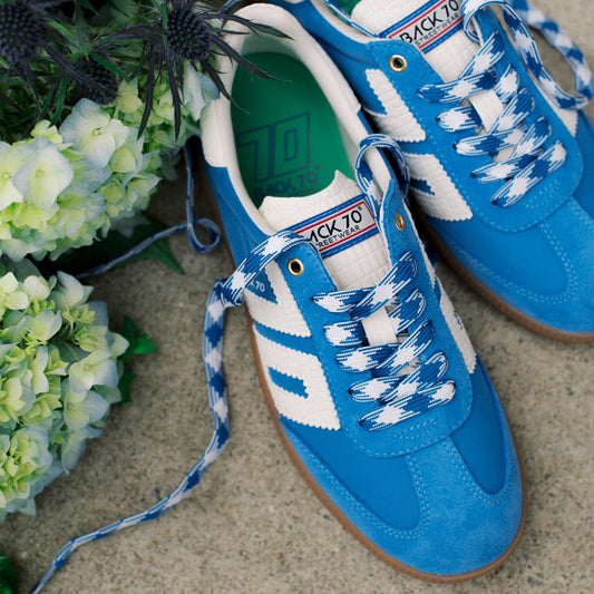 A pair of blue and white retro sneakers with blue-and-white checkered laces, styled on a textured concrete surface next to green hydrangea flowers.