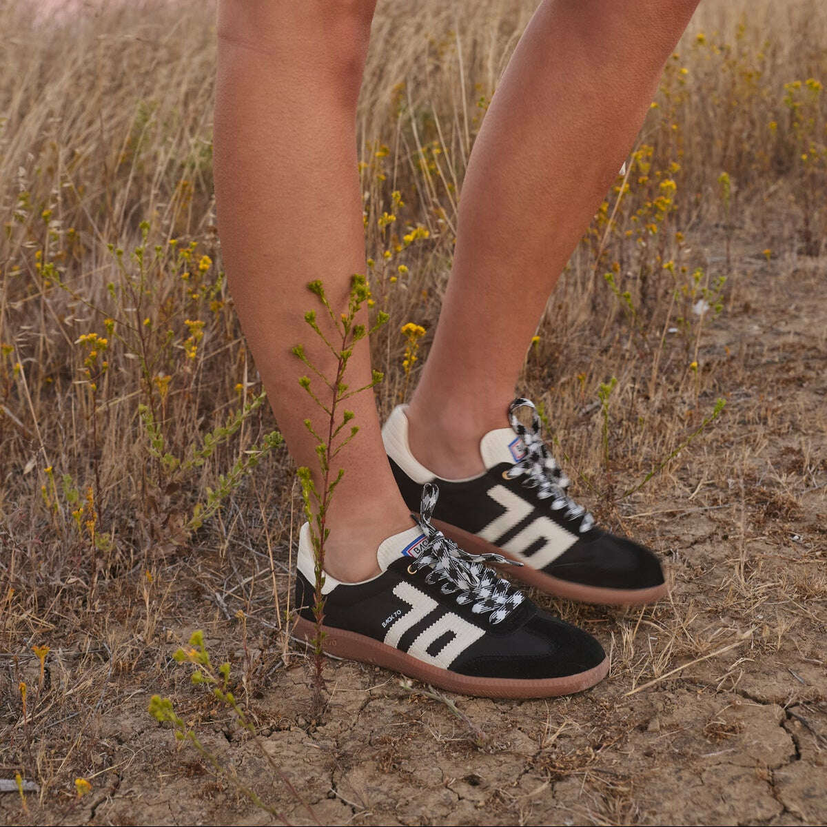 A person outdoors in a dry grassy field at sunset wearing black low-top sneakers with large white '76' lettering and white-and-black patterned laces, paired with a light cream skirt.