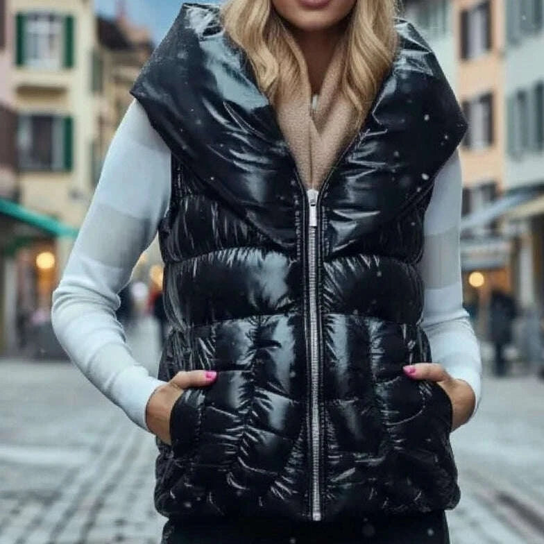 A woman wearing a shiny black quilted puffer vest over a white long-sleeve top and black pants, standing on a cobblestone street with buildings and snowy mountains in the background.