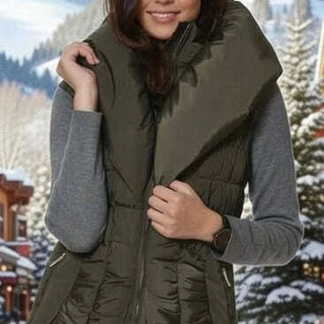 A woman wearing an olive green puffer vest over a gray long-sleeve shirt, standing in a snowy outdoor mountain setting.