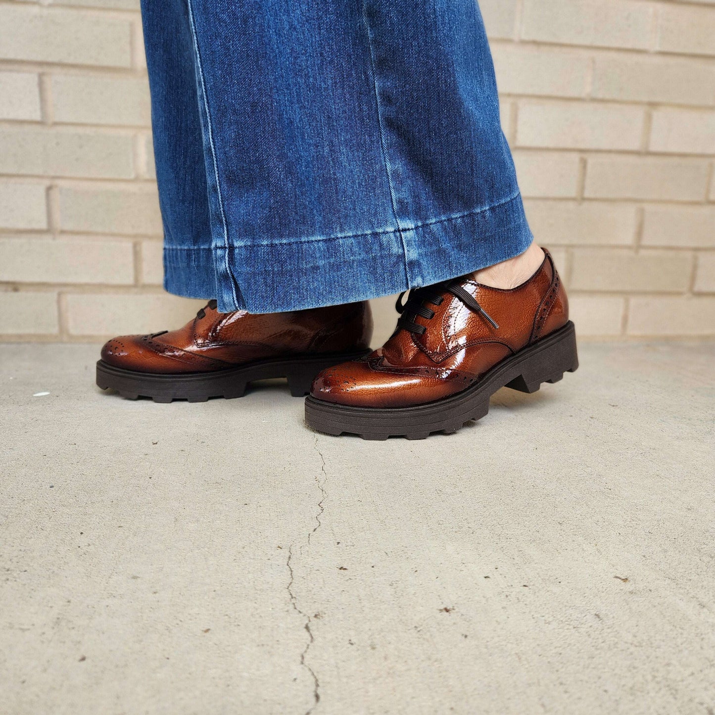 A person wearing brown glossy lace-up shoes with chunky black lug soles, paired with wide-leg blue jeans, standing on a concrete floor against a light brick wall.