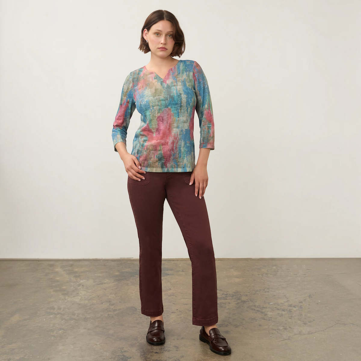 Plum Bottom FDJ notch neck top in a multi-colored abstract print with three-quarter sleeves, worn with brown trousers.