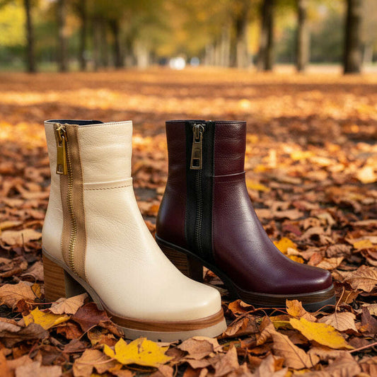 Two ankle boots displayed side by side on a textured gray surface in a retail store. The boot on the left is cream with a tan side panel and gold zipper pull.