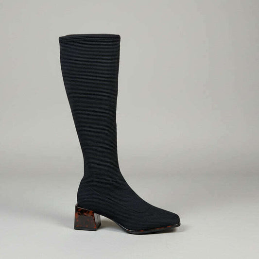 A single knee-high black ribbed boot with a square toe and a brown marbled block heel, photographed standing on a concrete floor against a light beige brick wall.
