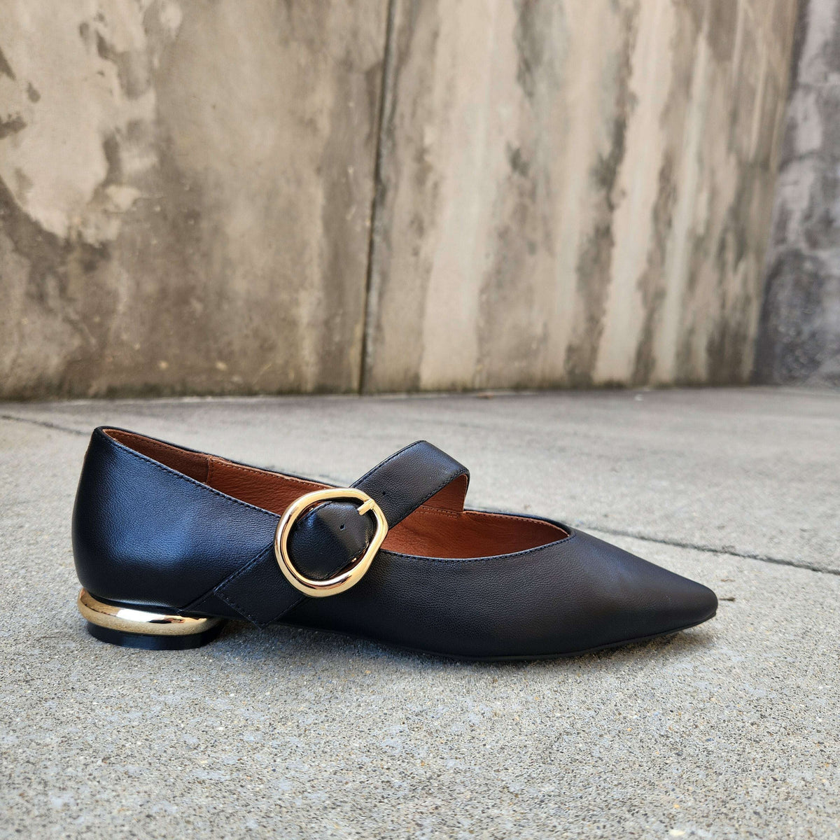 A single black leather Mary Jane flat shoe with a pointed toe, a gold-tone circular ring strap, and a low block heel with a gold-tone cap, resting on a concrete surface against a textured concrete wall.