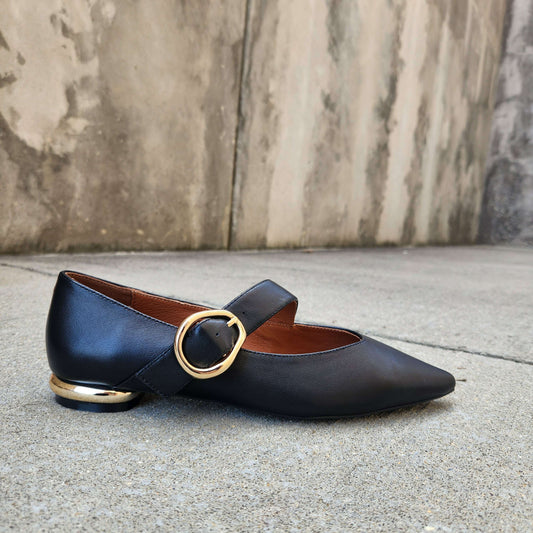 A single black leather Mary Jane flat shoe with a pointed toe, a gold-tone circular ring strap, and a low block heel with a gold-tone cap, resting on a concrete surface against a textured concrete wall.