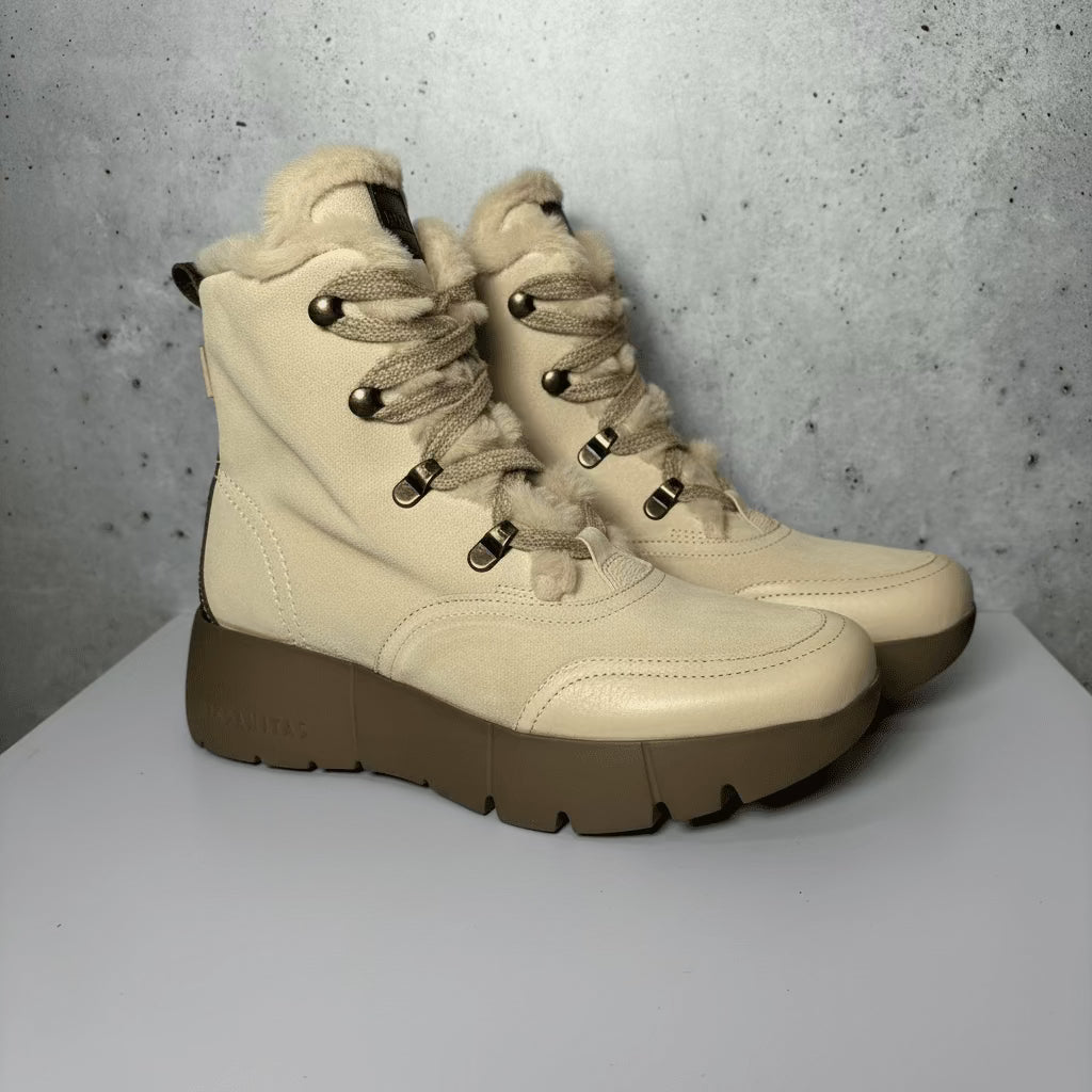 A single ankle-high platform winter boot in beige suede and tan rubber, featuring fuzzy brown laces, a plush beige lining, and a thick lug sole, photographed on a white platform against a gray textured wall.