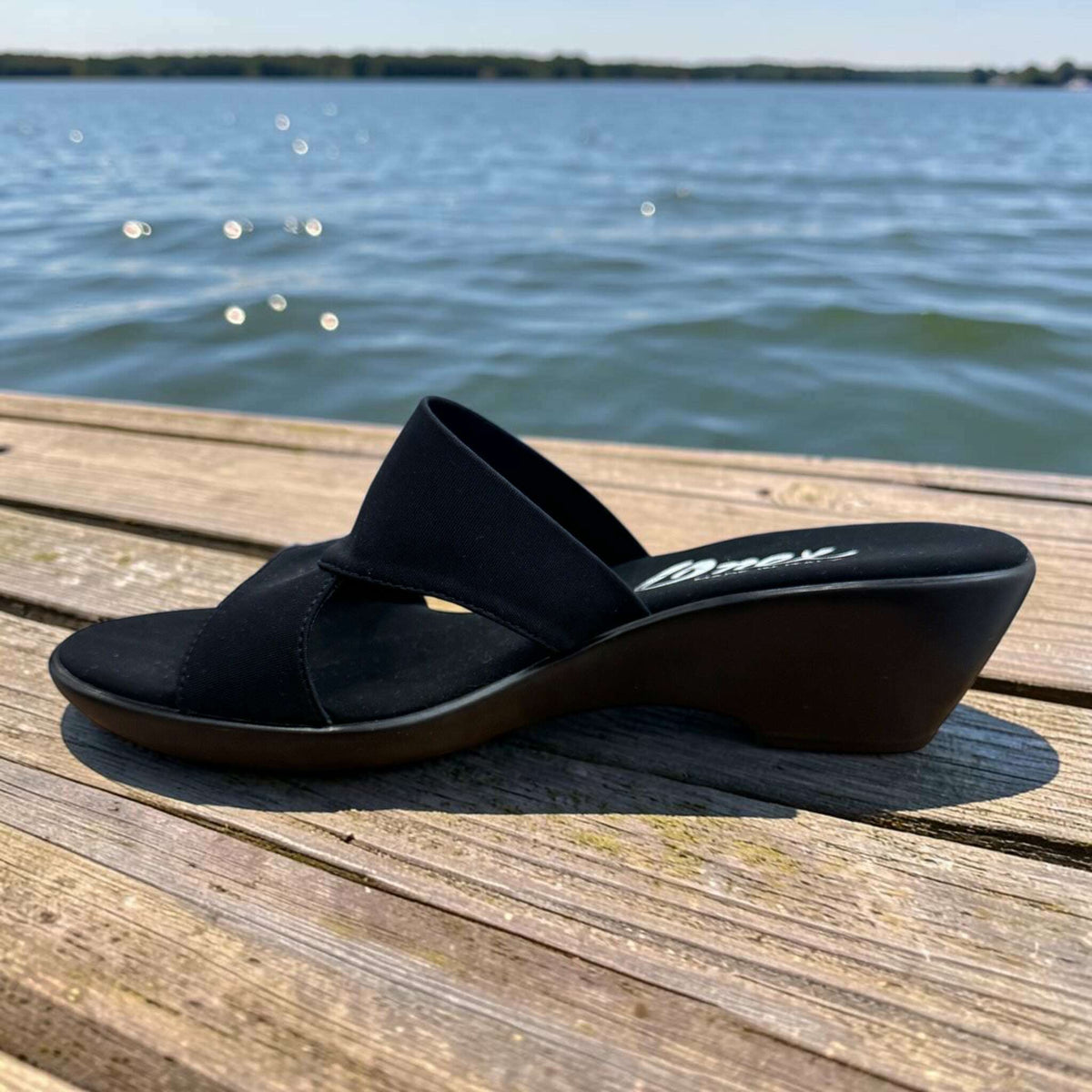 Plum Bottom ONEX Gilda-2 black slide sandal with crisscross straps and a 1.5-inch wedge heel on a wooden dock.