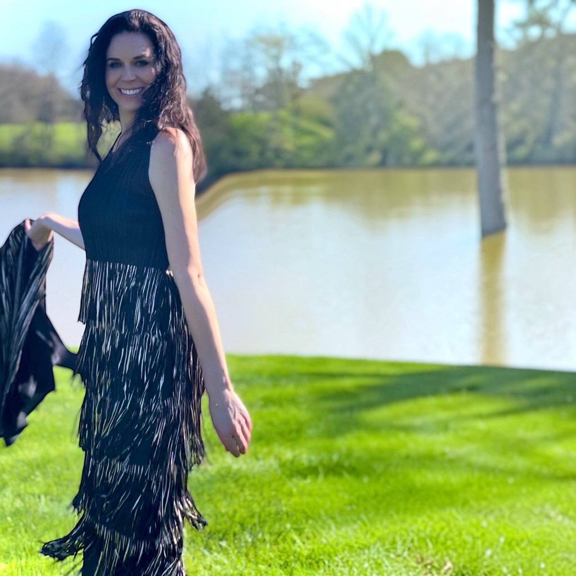 Plum Bottom Ellyndale black sleeveless pleated dress with gold foil fringe trim worn by a woman outdoors