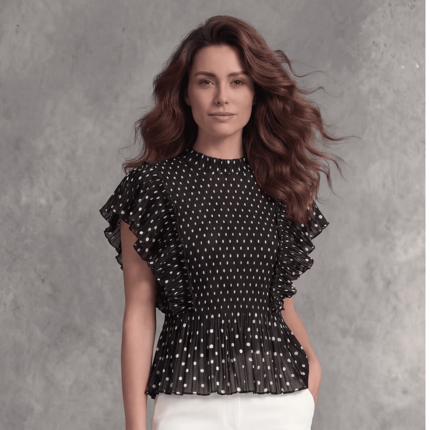 Plum Bottom black polka dot top with micro-pleating and cascading ruffle sleeves worn by a woman against a grey wall.