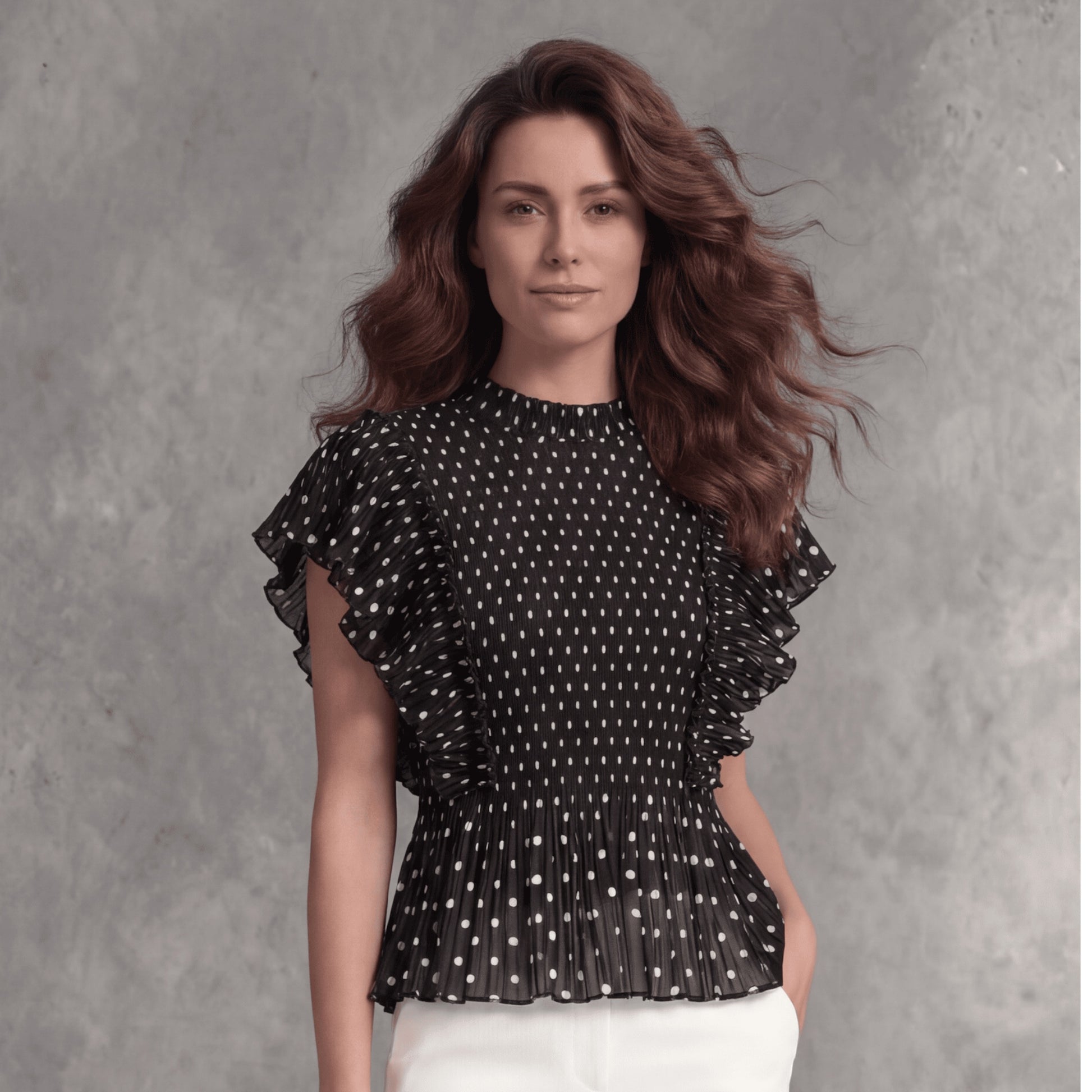 Plum Bottom black polka dot top with micro-pleating and cascading ruffle sleeves worn by a woman against a grey wall.