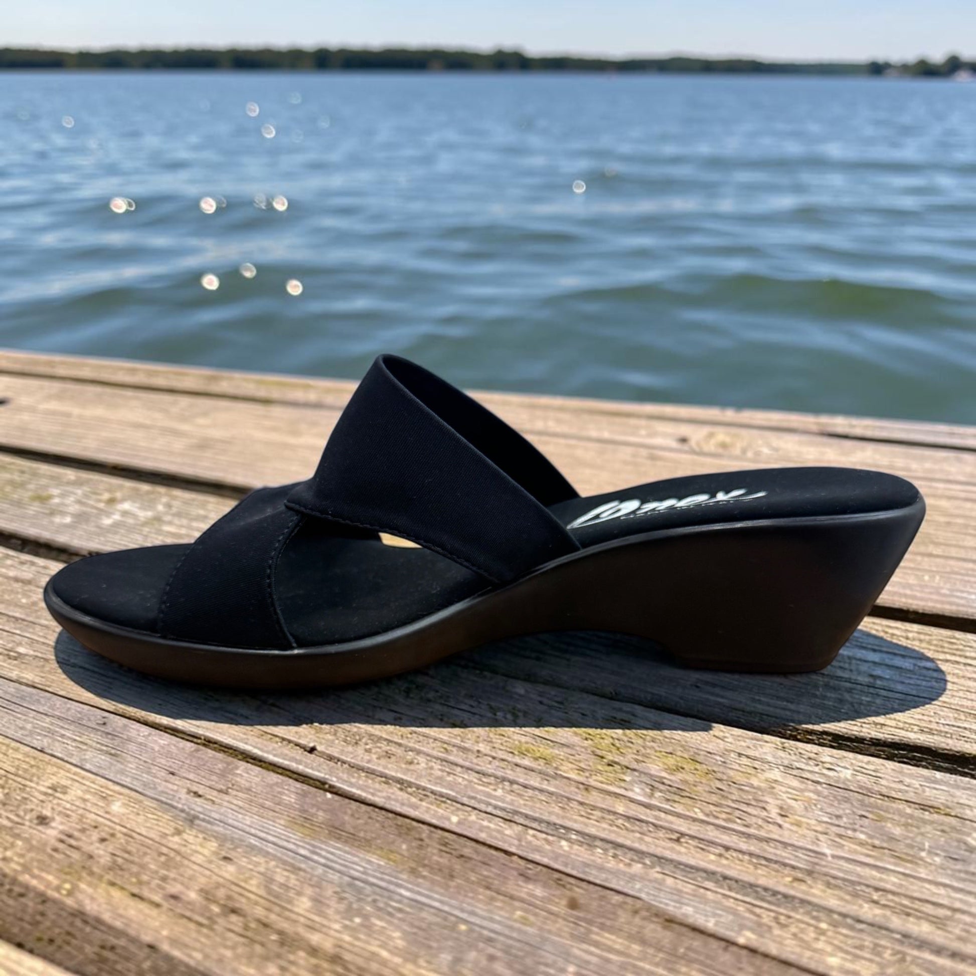 Plum Bottom ONEX Gilda-2 black slide sandal with crisscross straps and a 1.5-inch wedge heel on a wooden dock.