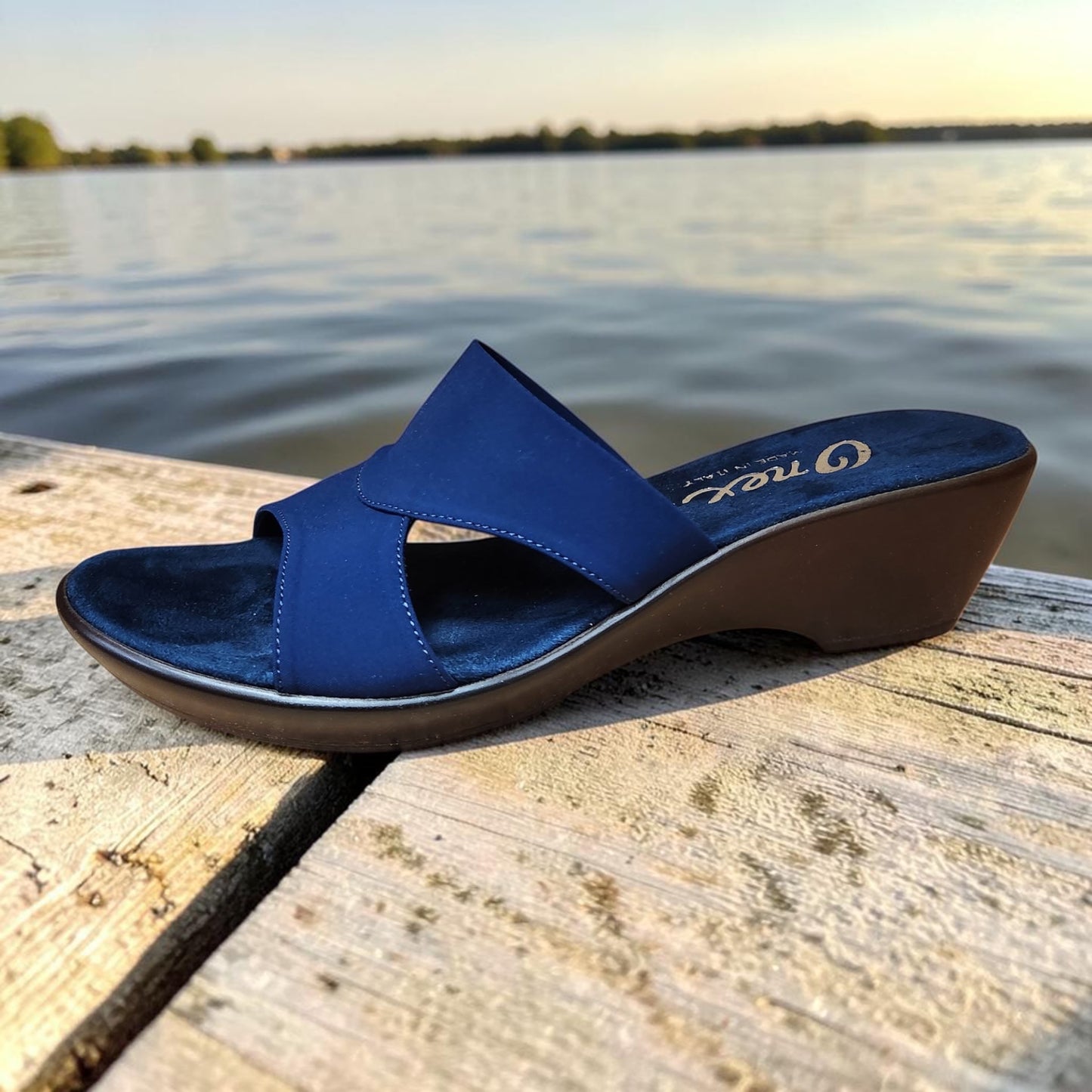 Plum Bottom ONEX Gilda-2 blue slide sandal with crisscross straps and a comfortable low wedge heel on a wooden dock.