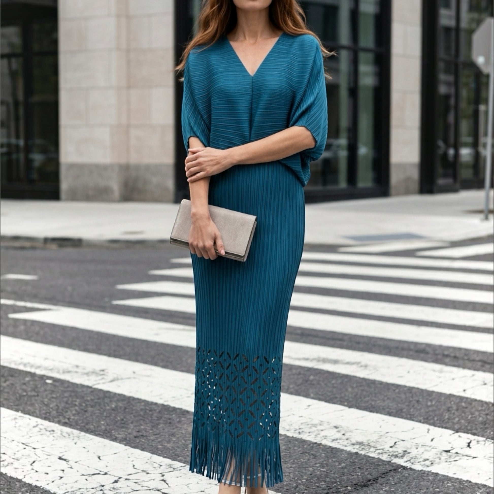 Plum Bottom teal pleated midi dress with waist cutout, laser-cut hem, and fringe detail worn on a city street.