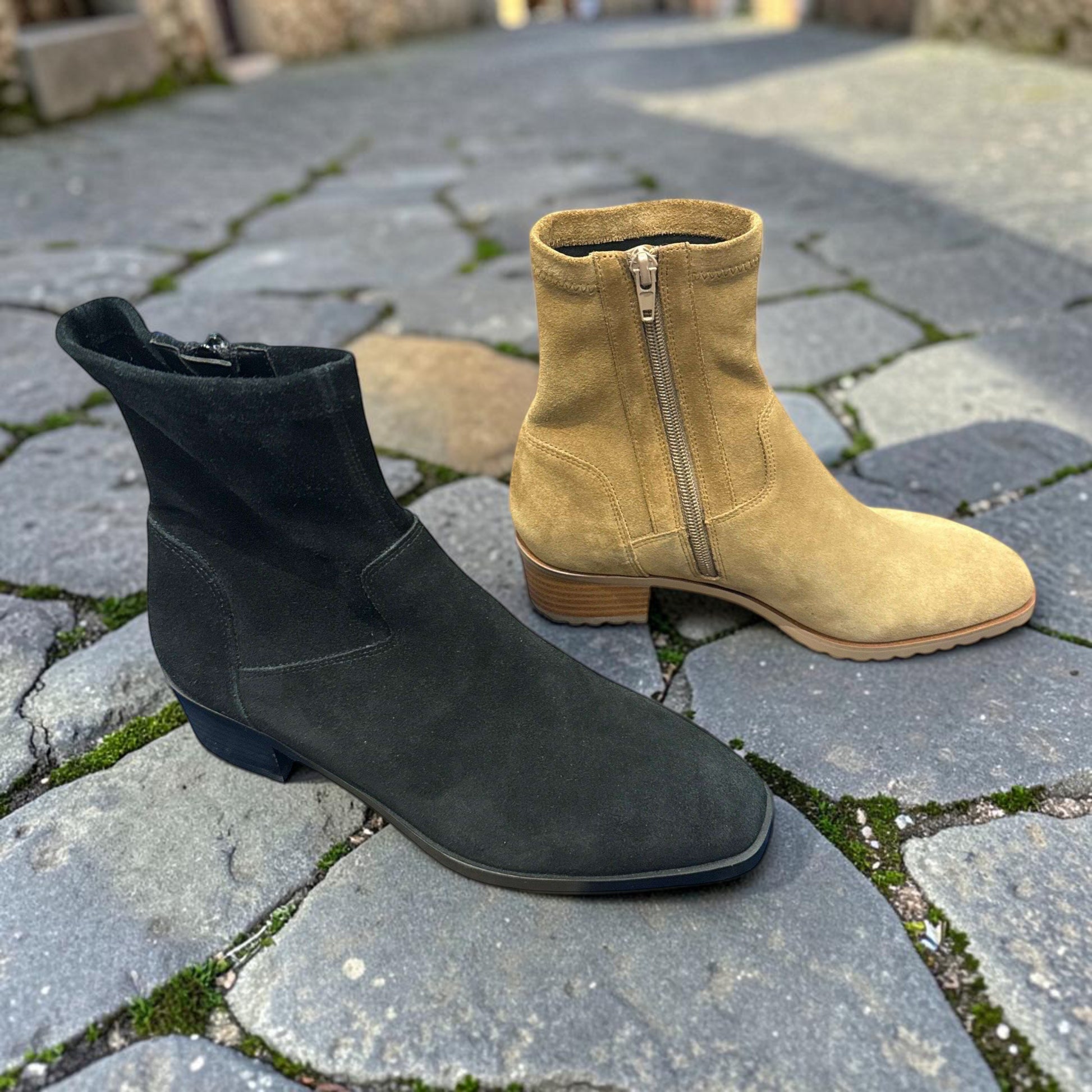 Plum Bottom VANELI Oleg suede booties in black and tan with side zippers and low block heels on a stone path.