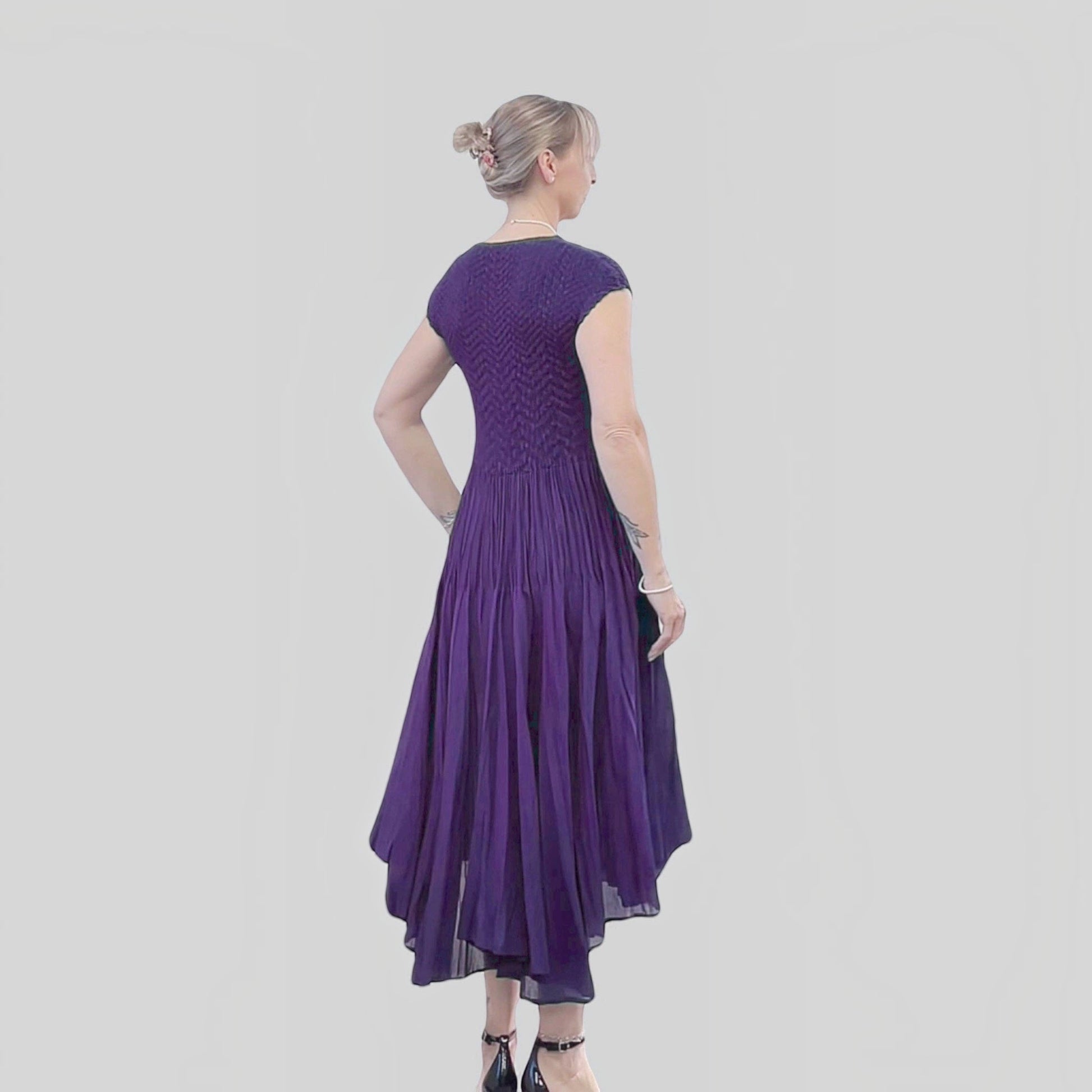 Plum Bottom violet pleated dress with cupsleeves worn by a woman, showing a flattering back view and flowy hem.
