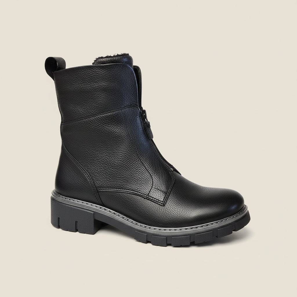 A single black leather ankle boot with a textured pebbled finish, side zipper, and chunky lug sole, shown against a corkboard background.
