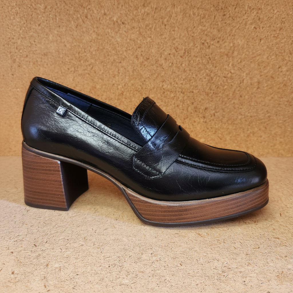 A single black leather penny loafer shoe with a chunky brown wooden block heel and platform sole, shown from a side profile against a corkboard background.