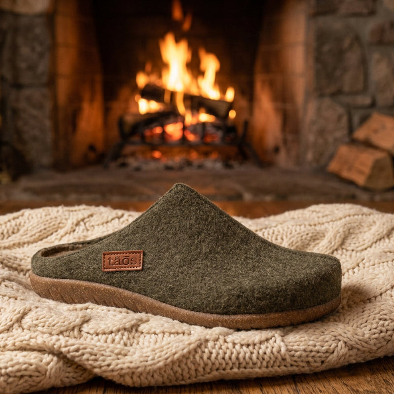 A single felt slip-on clog shoe in olive green with a brown sole, shown from a side profile on a white background. A small brown leather brand patch on the heel reads 'taos'.